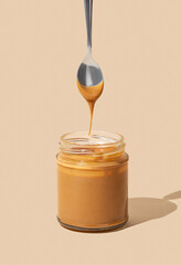 Peanut butter or caramel sauce dripping from spoon into glass jar on isolated beige background with shadow.