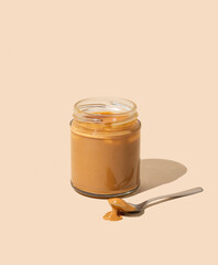 Peanut butter or caramel sauce in glass jar on isolated beige background with spoon and shadow.