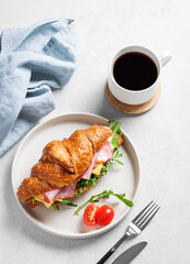 Croissant sandwich with ham, cheese, tomato  and arugula on a plate on a light background