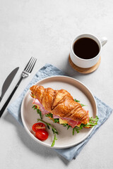 Croissant sandwich with ham, cheese, tomato  and arugula on a plate on a light background with a cup of  coffee.