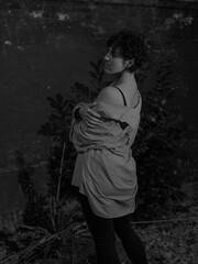 A young woman stands beneath dense foliage at night, holding a cigarette, dressed in casual attire,...