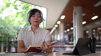 A university student in a white uniform sits at an outdoor study area, holding a book and wearing headphones around her neck, deeply engaged in reading.