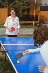 Obraz premium Two women engage in active game of ping pong at an outdoor table tennis setting. The area has green territory and wooden cabins, ideal summer outdoor leisure activities
