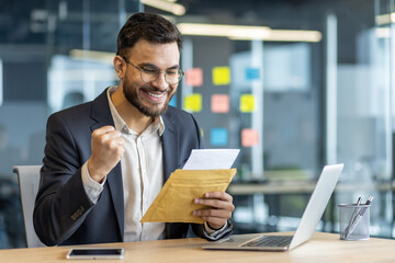 Successful businessman at workplace inside office with letter in hands. Man celebrates successful...