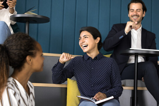 Group of professionals gathered in modern auditorium laughing, joking, sharing light-hearted moments, feel happy, enjoy friendly communication and friendship, focus on young Indian female, team leader