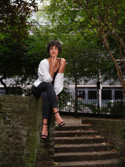 A young woman with curly hair sits gracefully on a brick wall in an urban park, wearing a black...