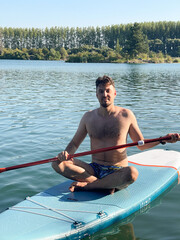 Man paddleboarding on calm lake water surrounded by greenery under a bright blue sky.