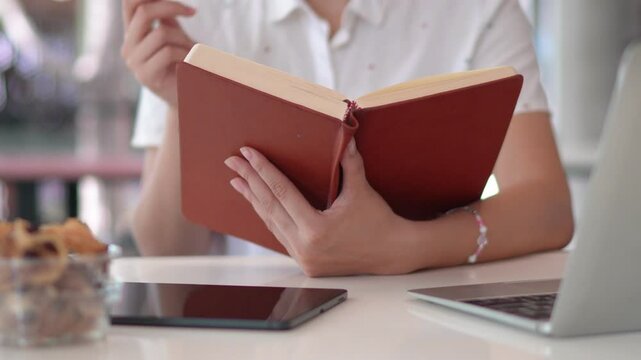 A university student in a white uniform reads a book while eating a snack in a casual study setting with a laptop nearby.