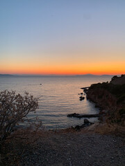 Coastal view at sunset with orange and blue gradient sky, calm sea, rugged cliffs, and sparse vegetation