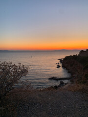 Coastal view at sunset with orange and blue gradient sky, calm sea, rugged cliffs, and sparse vegetation