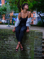 A young woman with curly hair sits on a moss-covered brick wall in an urban park, wearing a black...