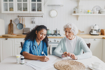 Indoor portrait of two people, young african american female doctor in blue uniform with stethoscope around her neck writing prescriptions for her elderly patient after detailed examination