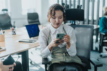 A young professional woman sits in an office chair, intently using her smart phone. The setting suggests a collaborative workspace with other colleagues engaged in work activities.