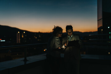 Two business people collaborate on a project at night using a digital tablet. The backdrop of a city skyline at dusk adds to the atmosphere of creativity and focus.