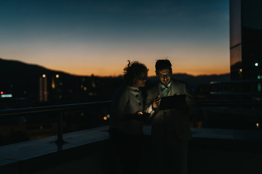 Two business people collaborate on a project at night using a digital tablet. The backdrop of a city skyline at dusk adds to the atmosphere of creativity and focus.