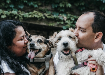 A smiling couple hugs and kisses their two dogs, who look happy with their tongues out. The background has green leaves, creating a warm and loving atmosphere.