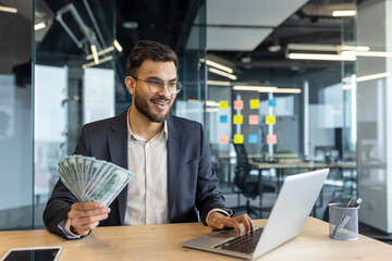 Successful joyful businessman working inside office with laptop. Man in business suit holding money...