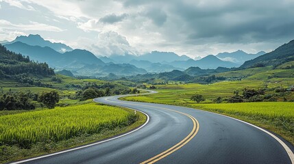 Naklejka premium Countryside roads nature concept. Winding road through lush green fields with mountains in the distance under a cloudy sky.
