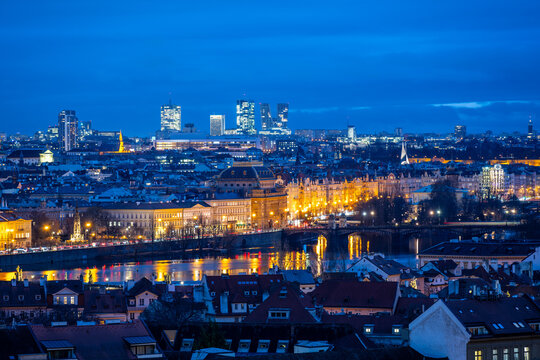 The National Theatre stands illuminated against the evening sky in Prague, with modern buildings of Pankrac rising in the background. Reflections shimmer in the water.