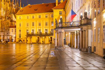 Fototapeta premium At dusk, the entrance door of Prague Castle showcases its impressive architecture, with a balcony adorned by statues. The cobblestones reflect the warm glow of the evening lights.