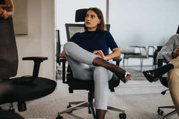 A thoughtful businesswoman sits in an office chair during a team meeting, reflecting on the discussion at hand. The modern office environment suggests a professional and collaborative workspace.