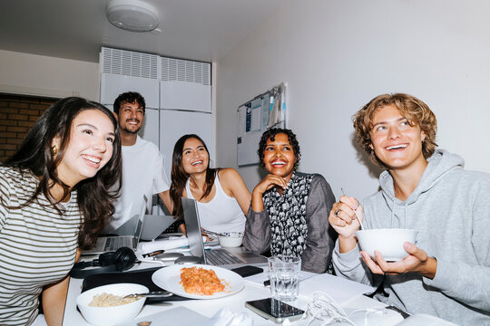 Happy multiracial roommates looking away while sitting at desk and studying in college dorm room