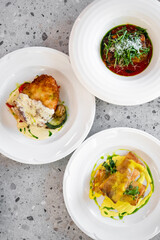 Three elegant plates of gourmet dishes are arranged on a textured surface. One features a baked entree with creamy sauce, while the other two showcase vibrant sauces and garnishes
