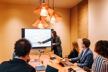 Female entrepreneur explaining graph diagram to colleagues on projection screen in board meeting at office