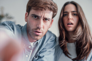 Obraz premium Confused couple touching camera clicking screen thinking worried doubting if click. Puzzled perplexed young two people with distrustful face unbelieving suspicious looking at home on sofa