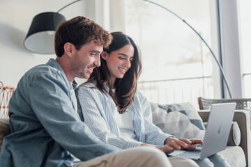 Happy millennial couple sit relax on couch in living room watching video on laptop together, smiling young husband and wife rest on sofa at home browsing Internet using modern computer device.