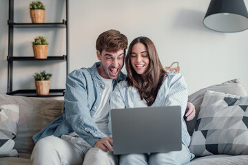 Happy emotional positive young family couple woman man looking at computer screen, celebrating online lottery giveaway betting auction win, feeling overjoyed together at home, internet success concept