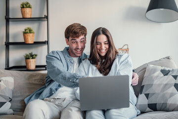 Happy millennial couple sit relax on couch in living room watching video on laptop together,...