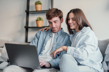 Happy millennial couple sit relax on couch in living room watching video on laptop together, smiling young husband and wife rest on sofa at home browsing Internet using modern computer device.
