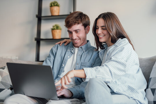 Happy millennial couple sit relax on couch in living room watching video on laptop together, smiling young husband and wife rest on sofa at home browsing Internet using modern computer device.
