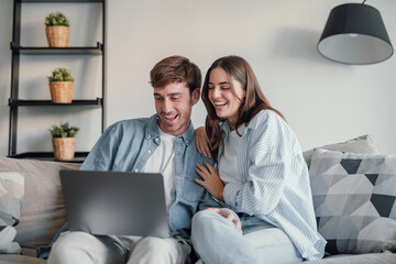 Happy millennial couple smiling looking at laptop screen making video call, young man and woman customers using computer applications for online shopping or booking sitting on sofa embracing.