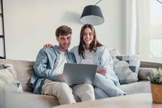 Happy millennial couple smiling looking at laptop screen making video call, young man and woman customers using computer applications for online shopping or booking sitting on sofa embracing.