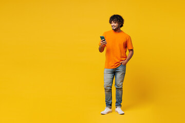 Full body young smiling happy Indian man he wears orange t-shirt casual clothes hold in hand use mobile cell phone chat online isolated on plain yellow background studio portrait. Lifestyle concept.