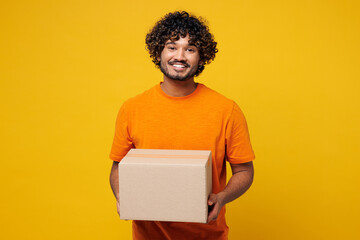 Fototapeta premium Young smiling cheerful fun cool happy Indian man he wear orange t-shirt casual clothes hold blank cardboard box looking camera isolated on plain yellow background studio portrait. Lifestyle concept.