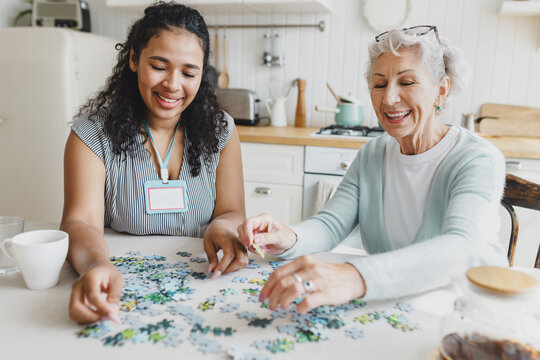 Indoor portrait of young black female volunteer and Caucasian old lady collecting puzzle together at kitchen table, having fun, talking and drinking tea. Social support for lonely elderly people