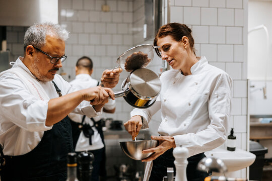 Male chef pouring ingredient in bowl held by female colleague at restaurant kitchen