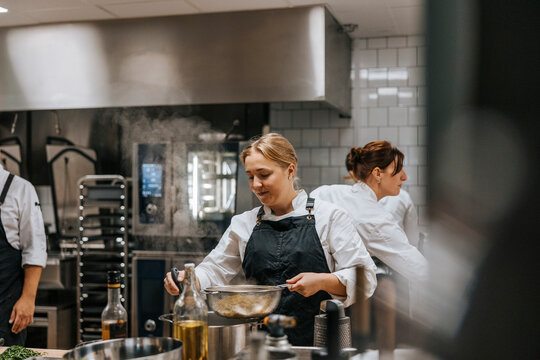Smiling young female chef straining Tagliatelle pasta in colander while working at restaurant kitchen