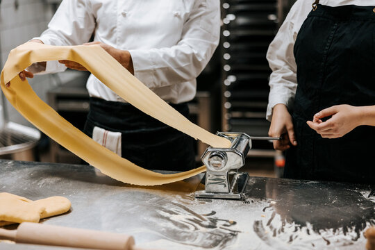Midsection of male chef stretching dough in pasta maker while standing with female colleague near kitchen counter at res