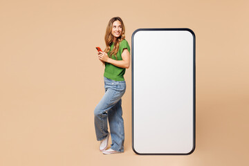 Full body side view happy young woman wear green t-shirt casual clothes big huge blank screen mobile cell phone with workspace area using smartphone isolated on plain pastel light beige background.