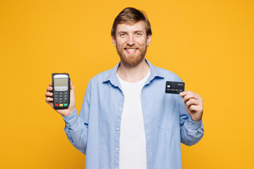 Young happy man wear blue shirt t-shirt casual clothes hold wireless modern bank payment terminal to process acquire credit card isolated on plain yellow orange background studio. Lifestyle concept.