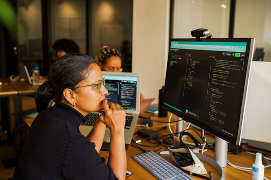 Thoughtful female coder looking at desktop PC while sitting in startup office
