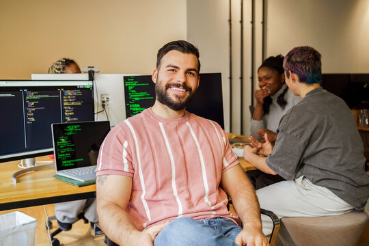 Portrait of smiling male coding professional sitting near desk with colleagues at office startup