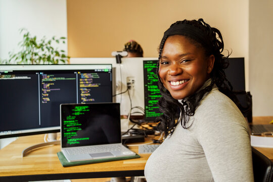 Portrait of happy young female It professional sitting near desktop PC and laptop in tech office