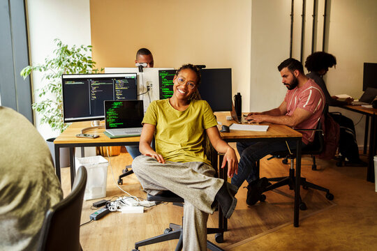 Portrait of happy female programmer sitting on chair in front of desktop PC at tech startup office
