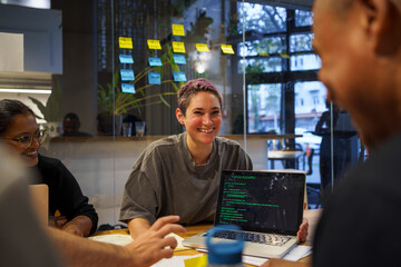 Happy non-binary programmer sitting with coded laptop in meeting room at office