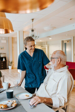 Smiling female healthcare worker talking with elderly male sitting at dining table in care home